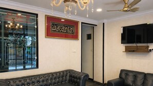 A room with two black leather sofas, a chandelier, a ceiling fan, a mounted TV, and a framed Arabic calligraphy piece on the wall. - Luciya The Boutique Hotel (Bengaluru)