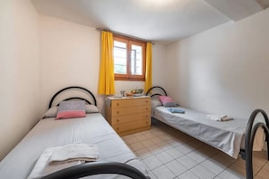 A small, clean room with two single beds, a wooden dresser, and a window with yellow curtains. - Beachside Apartment for a Relaxing Holiday (Olbia)