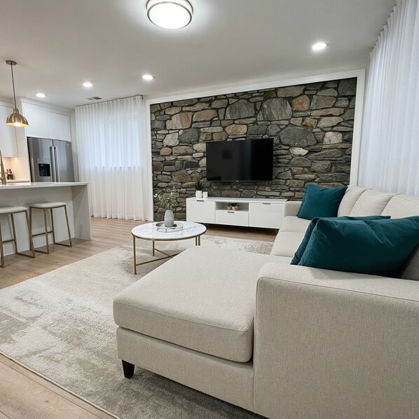 A modern living room with a stone wall, a flat-screen TV, a sofa with teal cushions, and a small round coffee table. - Luxury 1BR located in Prime Yonkers Area (Yonkers)