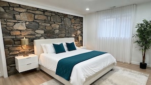 Ein Schlafzimmer mit Steinwand, einem Bett mit weißer Bettwäsche und Türkis-Akzenten, einem Nachttisch mit Lampe und einer Topfpflanze.