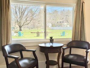A room with two leather chairs and a small table, a window with a view of a lawn and a house, and curtains on either side of the window. - Gallagher`s Travels Room 10 (Queen Bed) (Bar Harbor)