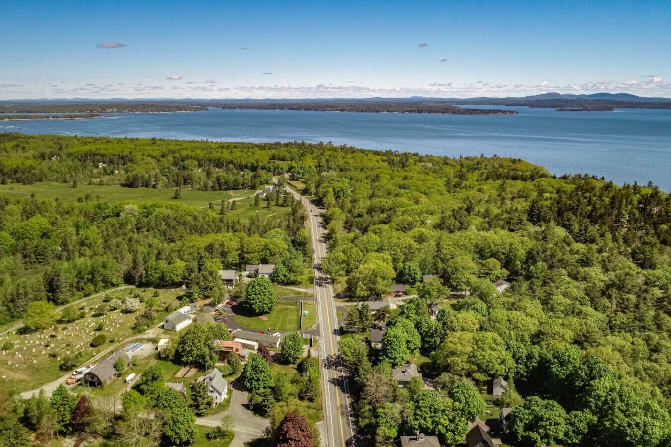 An aerial view of a residential area with a road running through it, surrounded by dense greenery and a large body of water in the background.
