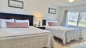 A hotel room with two beds, a nightstand with a lamp, a window with a view, and framed pictures on the wall. - Gallagher`s Travels Room 7 (Double Queen) (Bar Harbor)