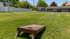 A backyard with a cornhole game set up on a well-maintained lawn, a white picket fence, and a small building in the background. - Gallagher`s Travels Room 7 (Double Queen) (Bar Harbor)