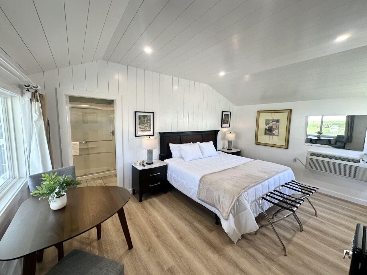 A modern bedroom with a large bed, bedside tables with lamps, a small table with a plant, a chair, and a window with curtains.