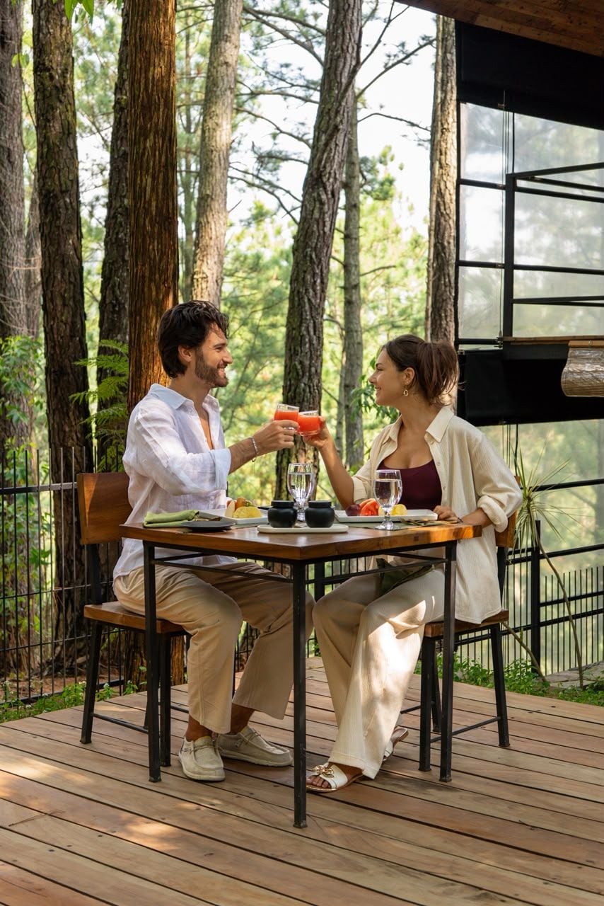 Two people seated at an outdoor table with a glass of drink, surrounded by trees.