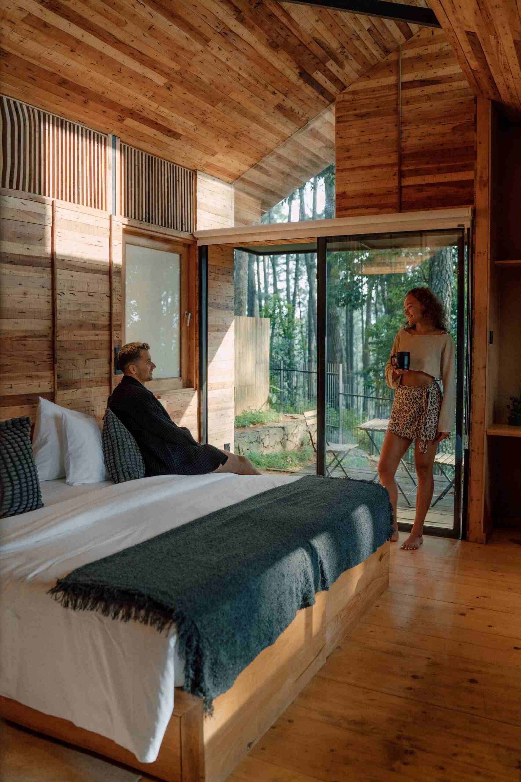 A wooden cabin interior with a bed, a person sitting on the bed, and another person standing by a sliding glass door.