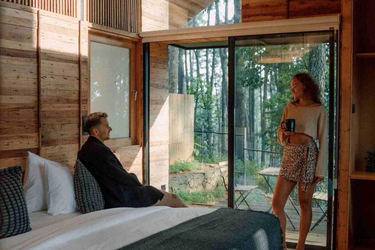 A wooden cabin interior with a bed, a person sitting on the bed, and another person standing by a sliding glass door.