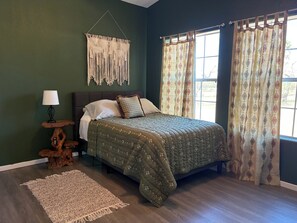A bedroom with a bed, a nightstand, a lamp, a macrame wall hanging, and a window with curtains.