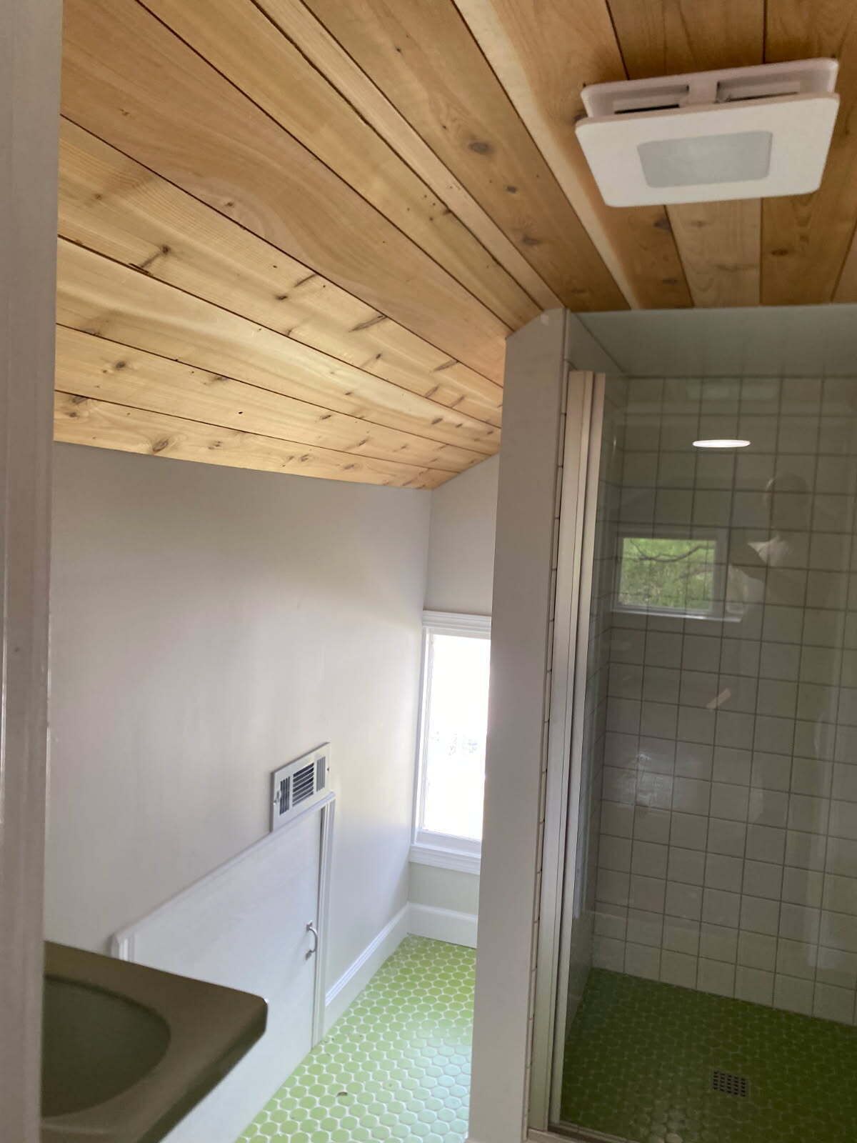 A bathroom with a wooden ceiling, a shower with green tiles, a window, and a toilet.