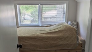 A bedroom with a sloped ceiling, a window with blinds, a bed with a beige blanket, a bedside table with a lamp, and a carpeted floor. - Charming Stone Cottage On Lake James / Lone Tree Point (Angola)
