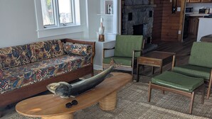 A living room with a patterned sofa, green chairs, a wooden coffee table, and a stone fireplace. - Charming Stone Cottage On Lake James / Lone Tree Point (Angola)