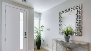 Un corridoio moderno con una porta bianca, una pianta in vaso, un tavolo consolle con uno specchio geometrico e un decoro parietale a forma di sole.