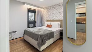 Un dormitorio moderno con cama, ventana con cortinas, un espejo y una pequeña kitchenette visible a través del espejo.