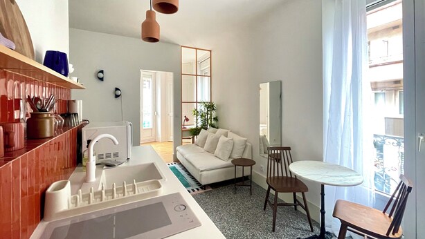 Une chambre d’hôtel moderne dotée d’une kitchenette, d’un canapé, d’une table à manger avec des chaises et d’une grande fenêtre avec des rideaux.
