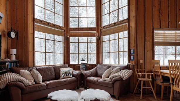 A cozy cabin interior with a brown leather sofa, two white furry ottomans, a wooden dining table with chairs, and large windows offering a view of snow-covered trees. - Alpine Fox by Aavalevi (Levi)