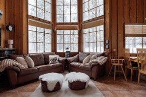 Interior acogedor de una cabaña con un sofá de cuero marrón, dos otomanas blancas y mullidas, una mesa de comedor de madera con sillas y ventanas amplias que ofrecen vistas de árboles cubiertos de nieve.