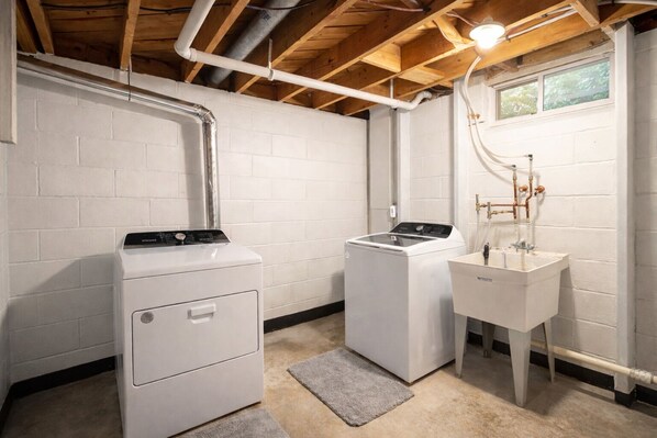 A laundry room with two white washers and a sink, wooden ceiling beams, and white brick walls. - Private Hot Tub • Bright Home Near Downtown (Oconomowoc)