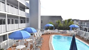 Un área de piscina en la azotea con sillones de descanso y sombrillas, rodeada por un edificio de varios pisos con balcones.