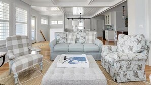 Un vaste salon avec un canapé bleu clair, un fauteuil à carreaux et un fauteuil à motifs. On y trouve également un ventilateur de plafond, un lustre et, au fond, un coin repas.
