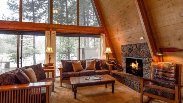 A cozy, log cabin-style room with a fireplace, wooden furniture, and large windows offering a view of the forest. -  Inviting Central Oregon Escape (Sunriver)