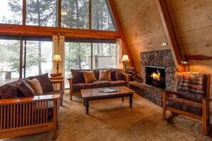 Une pièce chaleureuse, aménagée dans le style d’un chalet en bois, avec une cheminée, des meubles en bois et de grandes fenêtres donnant sur la forêt.
