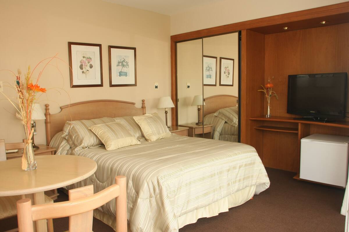 A hotel room with a bed, a desk, a chair, a television, and framed pictures on the wall.