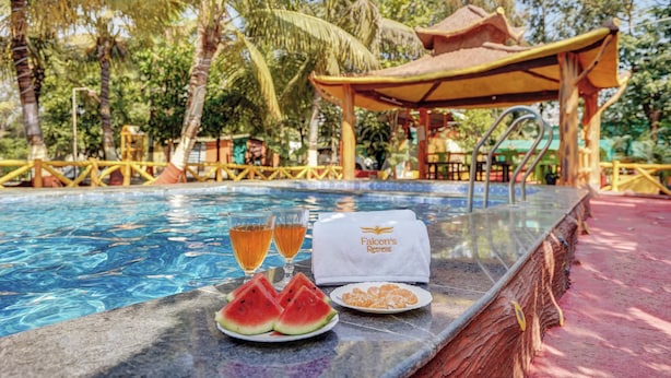 Escena junto a la piscina con una mesa de vidrio, dos vasos de una bebida, rebanadas de sandía y un plato de naranjas. También se ve una toalla con el logotipo de "Falcon's Retreat".
