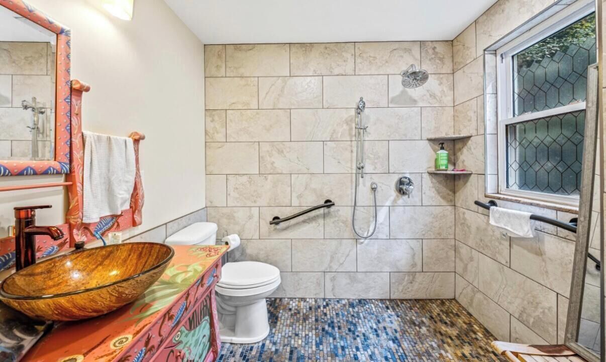 Une salle de bain avec un sol en mosaïque, un lavabo en forme de bol en bois, des toilettes, une douche et une fenêtre avec une grille en métal.
