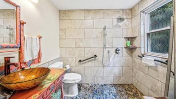 Une salle de bain avec un sol en mosaïque, un lavabo en forme de bol en bois, des toilettes, une douche et une fenêtre avec une grille en métal.
