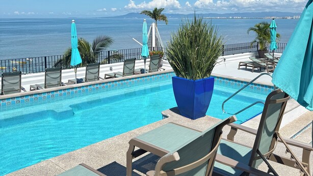 Zona de piscina con sillones de descanso y sombrillas, con vista al mar azul y montañas distantes.