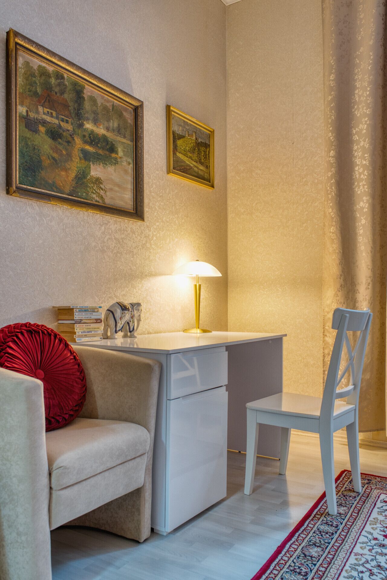 Ett mysigt rum med ett skrivbord, en stol och en röd kudde på en soffa. På väggen hänger två inramade tavlor och på skrivbordet står en lampa.