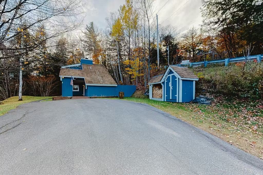 A wide paved driveway leads to a blue house and matching woodshed nestled in the trees.