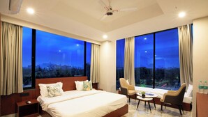A modern hotel room with a large bed, two chairs, a small table, and a city view. - Aarchh Regency (Noida)