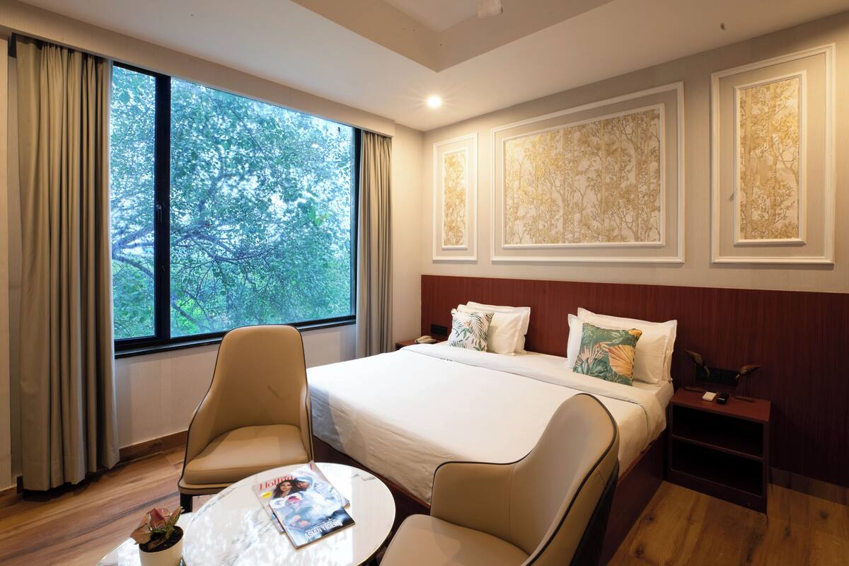 A hotel room with a large window, a bed with white linens, two armchairs, a small round table, and a nightstand with a lamp.