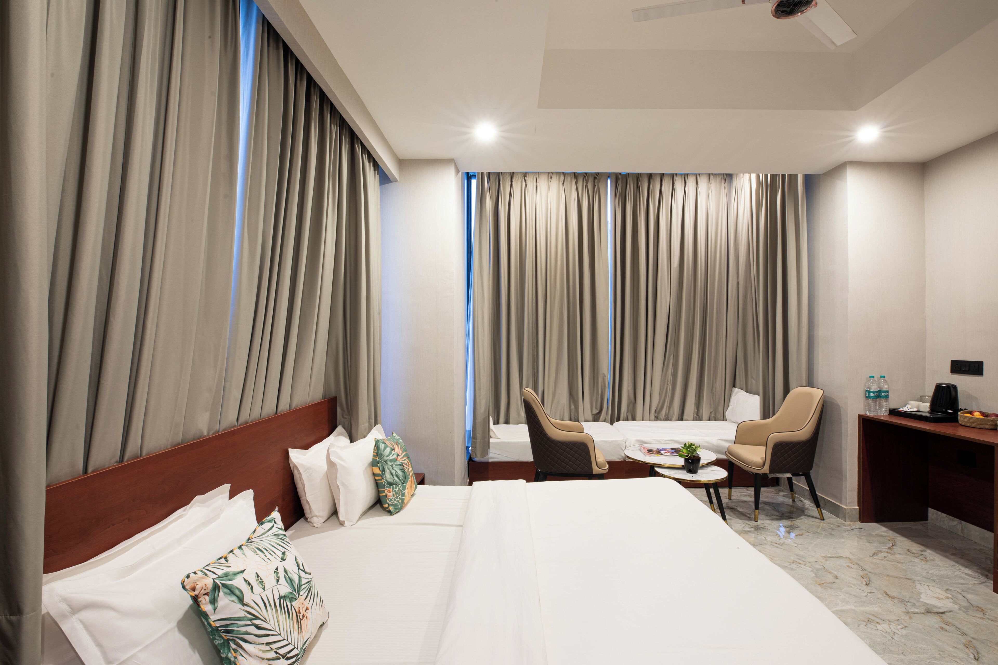 Een hotelkamer met een groot bed, een bureau met een stoel, een bijzettafeltje met een plant en gordijnen voor de ramen.