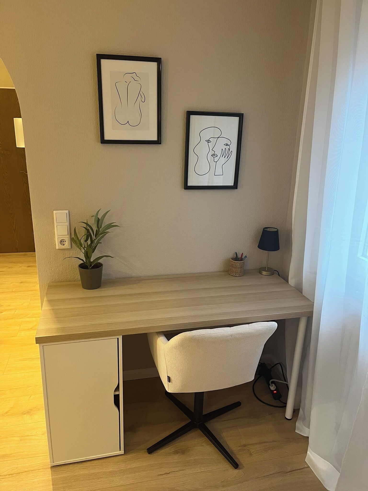 A modern workspace with a wooden desk, a white chair, a potted plant, and two framed artworks on the wall.