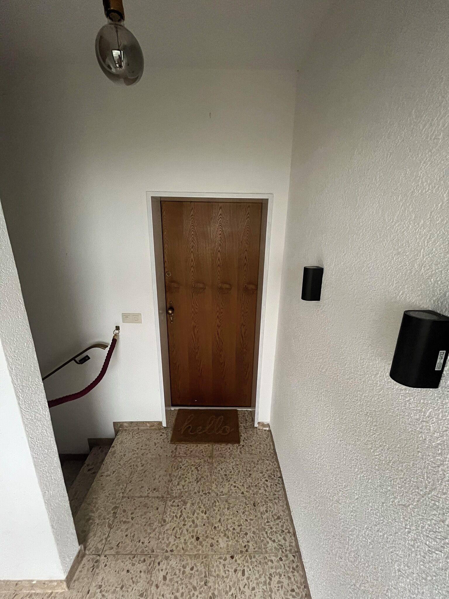 A hallway with a wooden door, a doormat, a wall-mounted device, and a red hose.