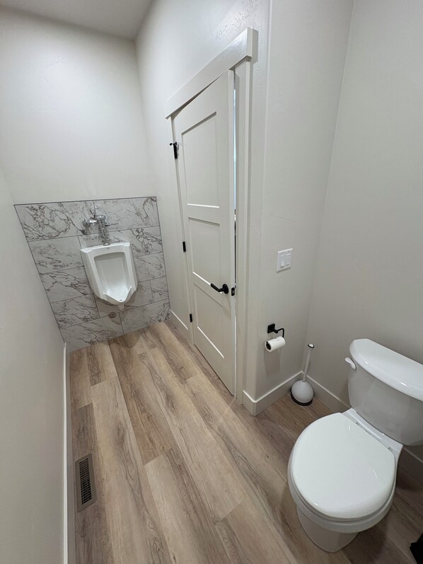 Ein Badezimmer mit einer Wand aus Marmorfliesen, Holzboden, Toilette, Urinal und einer Tür mit Griff.