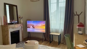 A living room with a fireplace, a television on a stand, a dining table with chairs, and a large window with curtains. - Maison Le Beaulieu (Vichy)