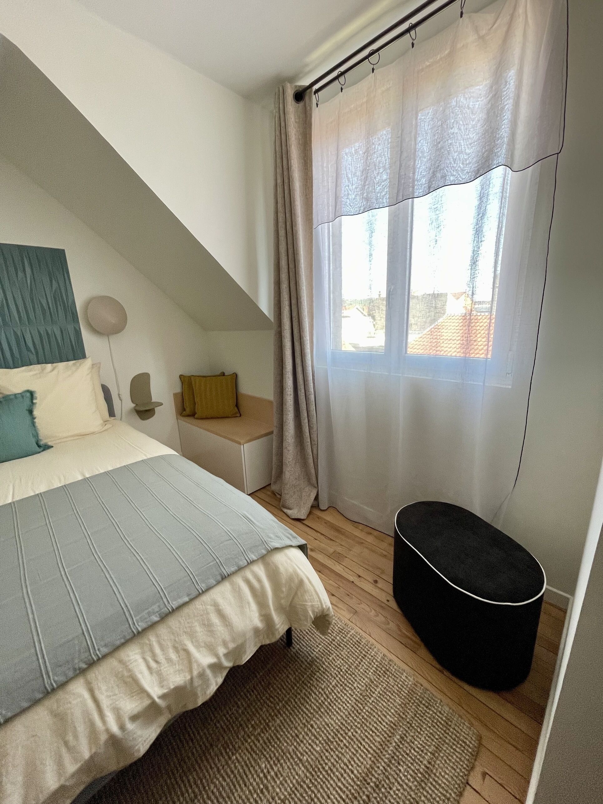 Un dormitorio con cama, mesita de noche pequeña, lámpara, ventana con cortinas y un reposapiés negro.
