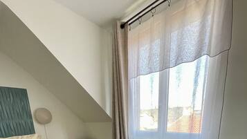 Un dormitorio con cama, mesita de noche pequeña, lámpara, ventana con cortinas y un reposapiés negro.