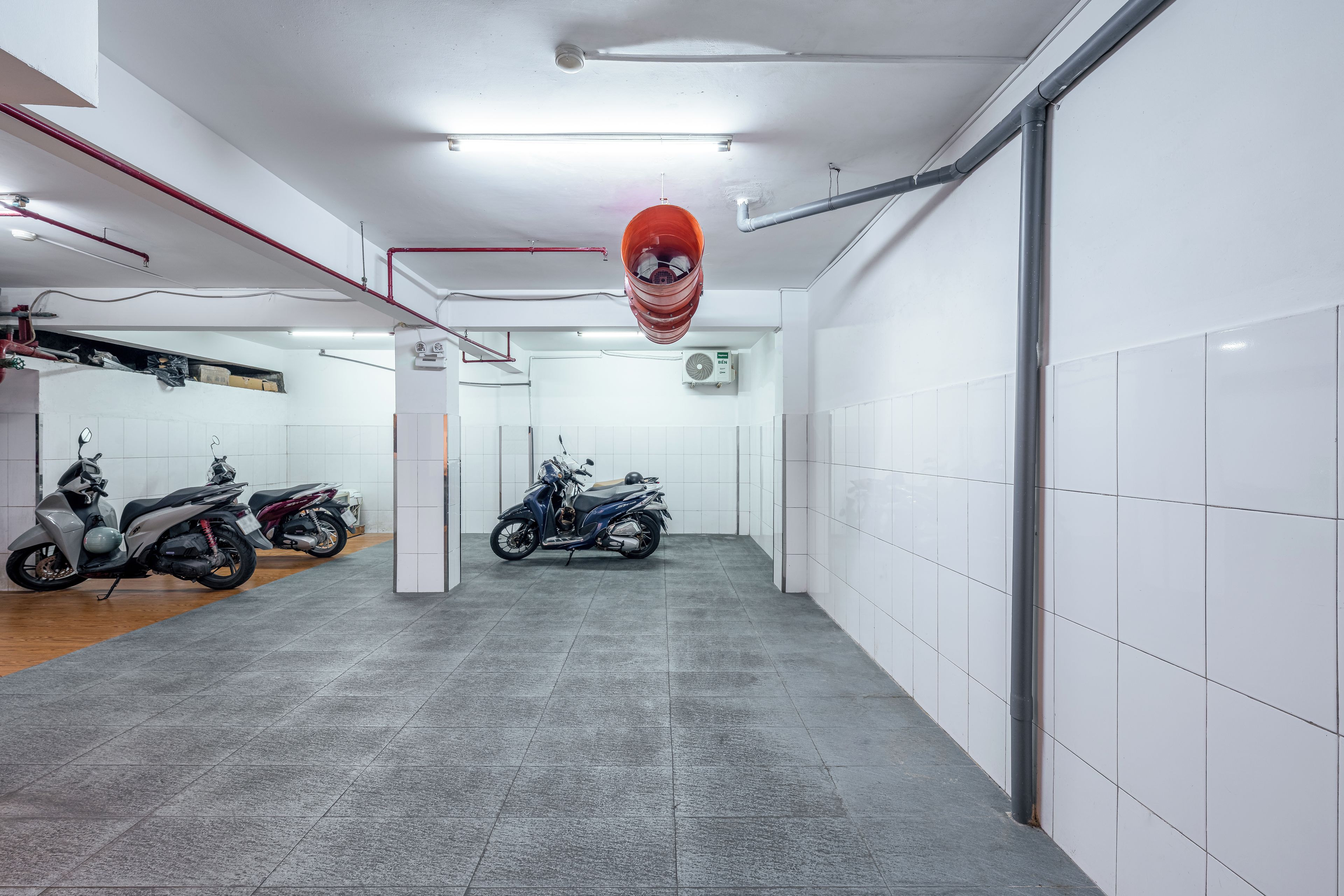 A clean, well-lit indoor space with tiled walls and a concrete floor, featuring two parked scooters and visible plumbing fixtures.