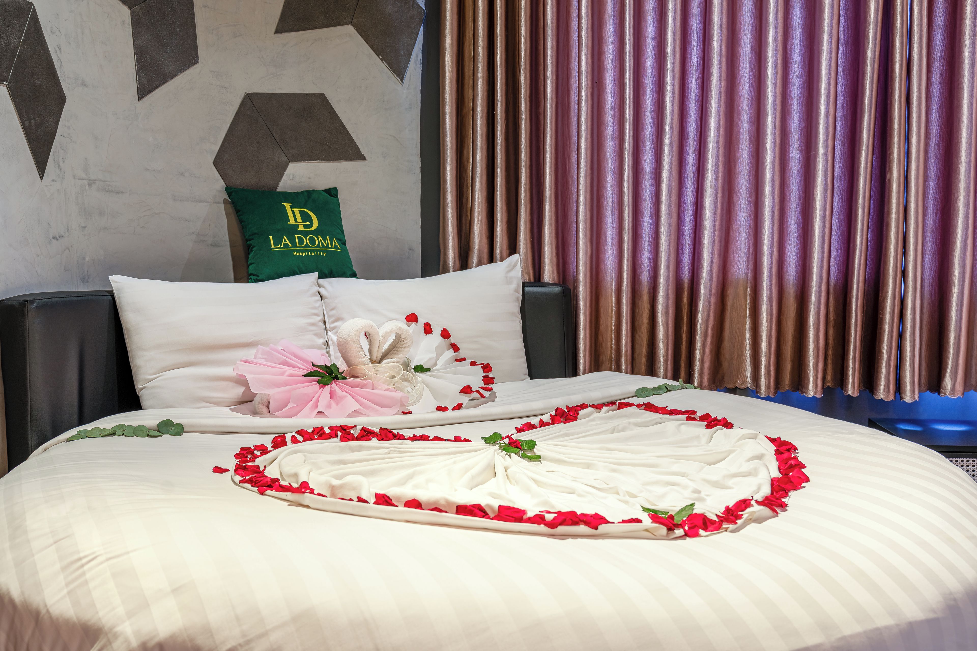 A hotel room with a bed, pillows, a green bag with 'ID LA DOMA' text, and a curtain.