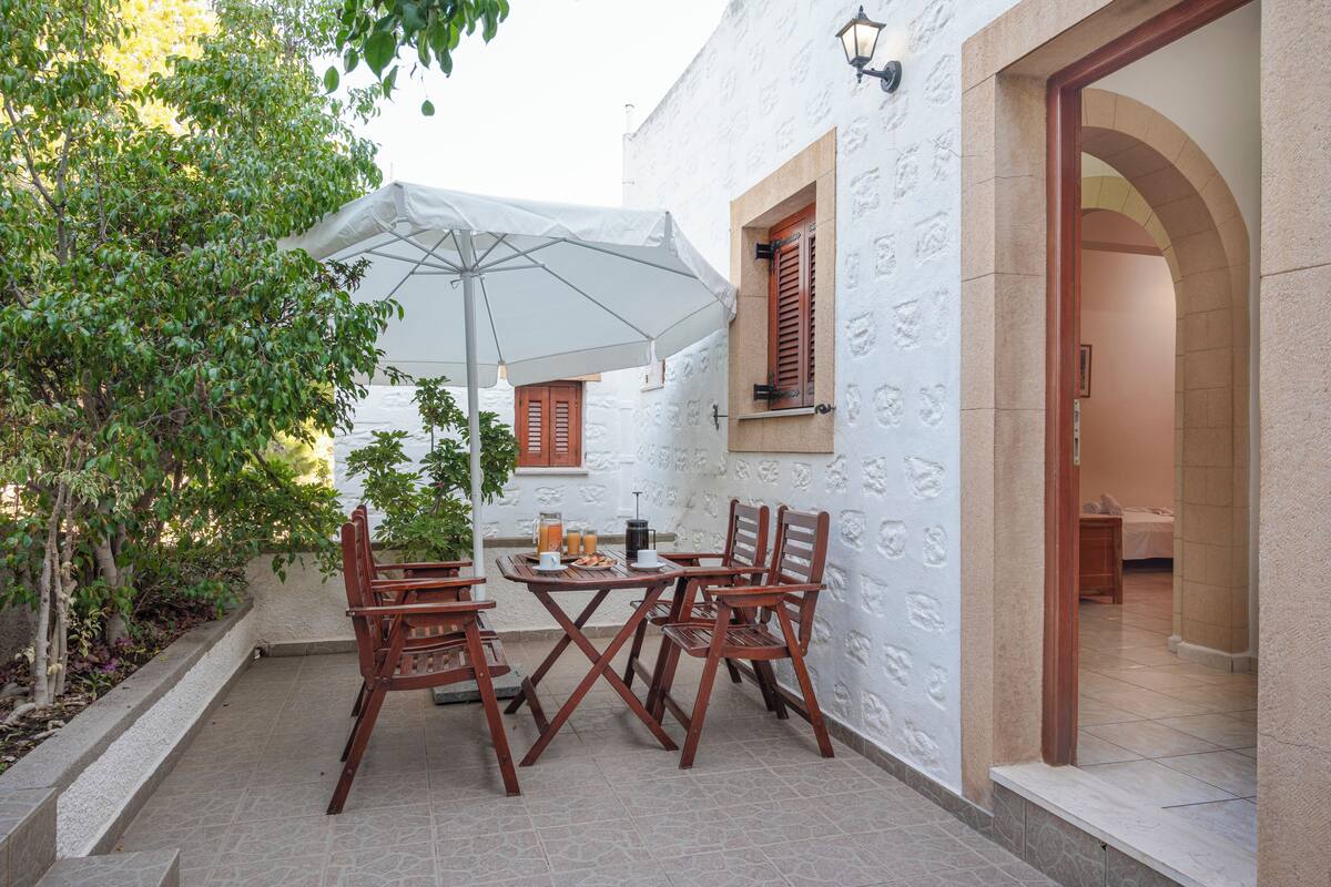 An outdoor patio with a table and chairs, a white umbrella, and a view of a building with a tiled floor and a doorway.