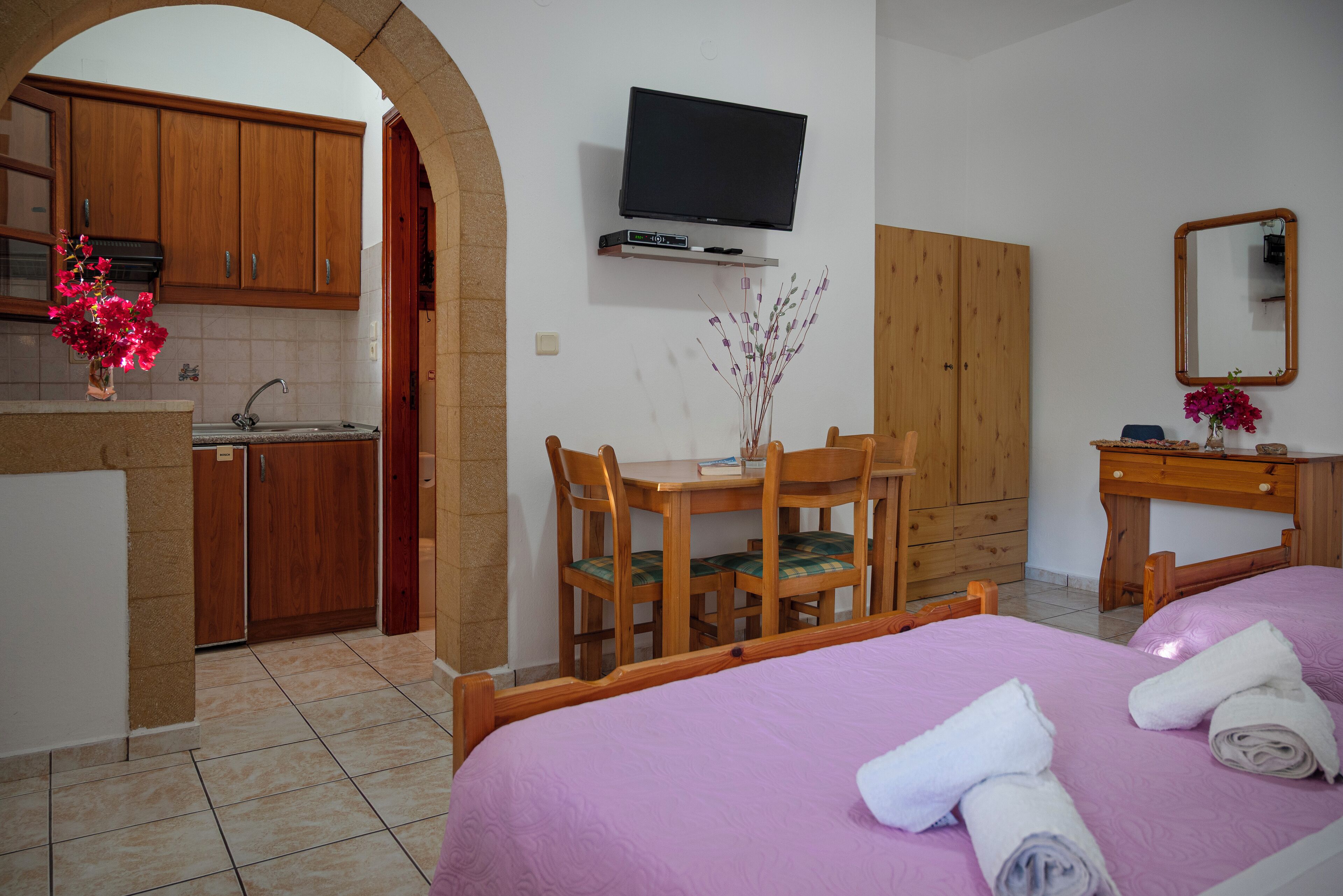 A hotel room with a bed, a desk with chairs, a television mounted on the wall, and a kitchen area visible through an archway.