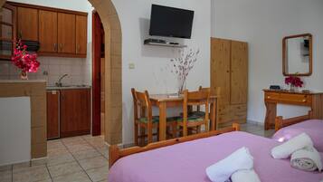 A hotel room with a bed, a desk with chairs, a television mounted on the wall, and a kitchen area visible through an archway.