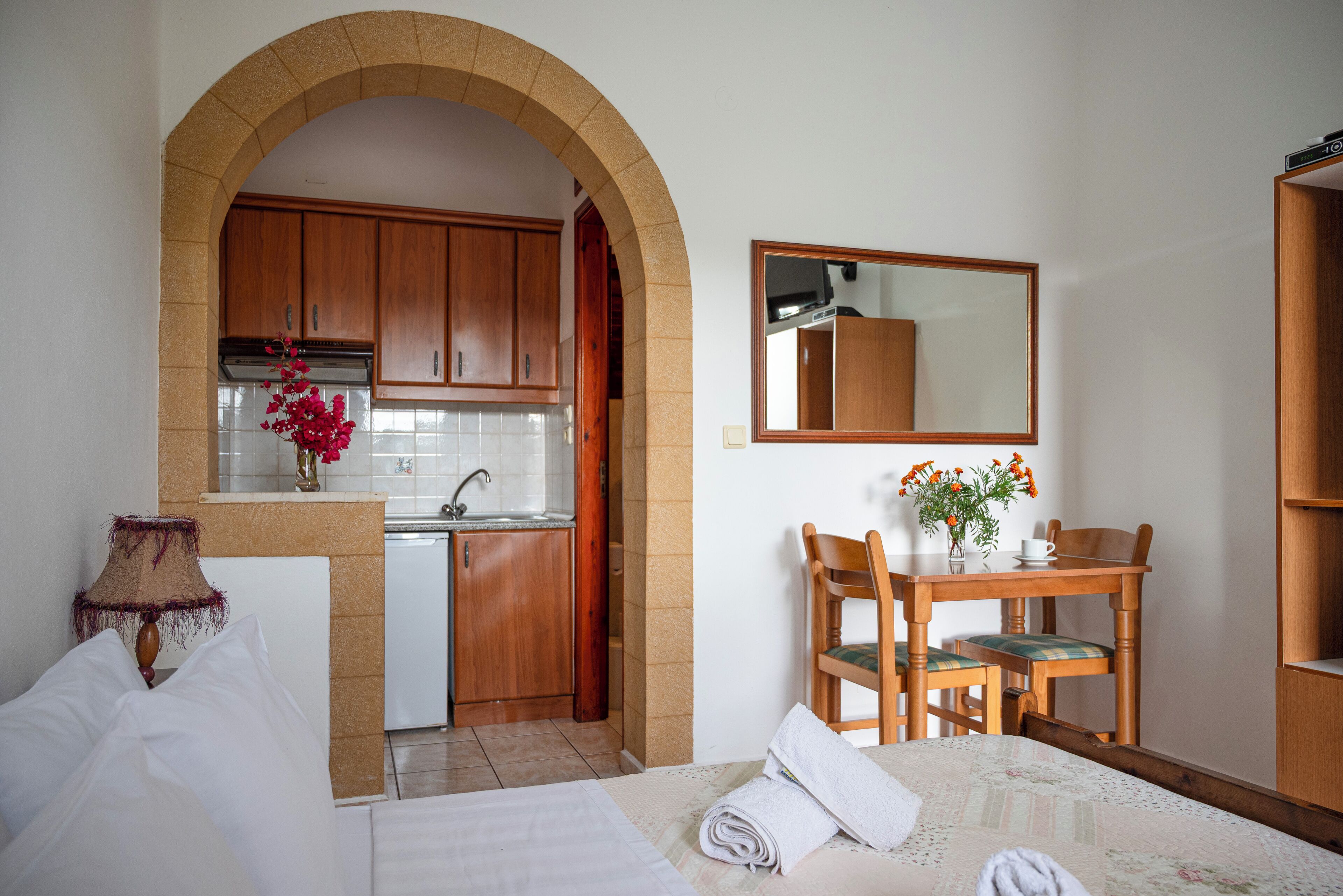 Habitación de hotel con cama, escritorio, silla, pequeña mesa, espejo y cocina con armarios de madera y un lavabo.