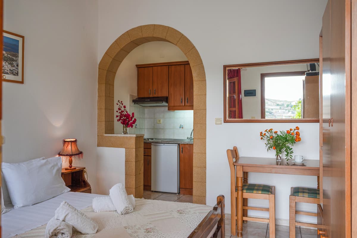 A bedroom with a bed, a bedside table with a lamp, a small kitchenette, a wooden desk with a chair, and a mirror.