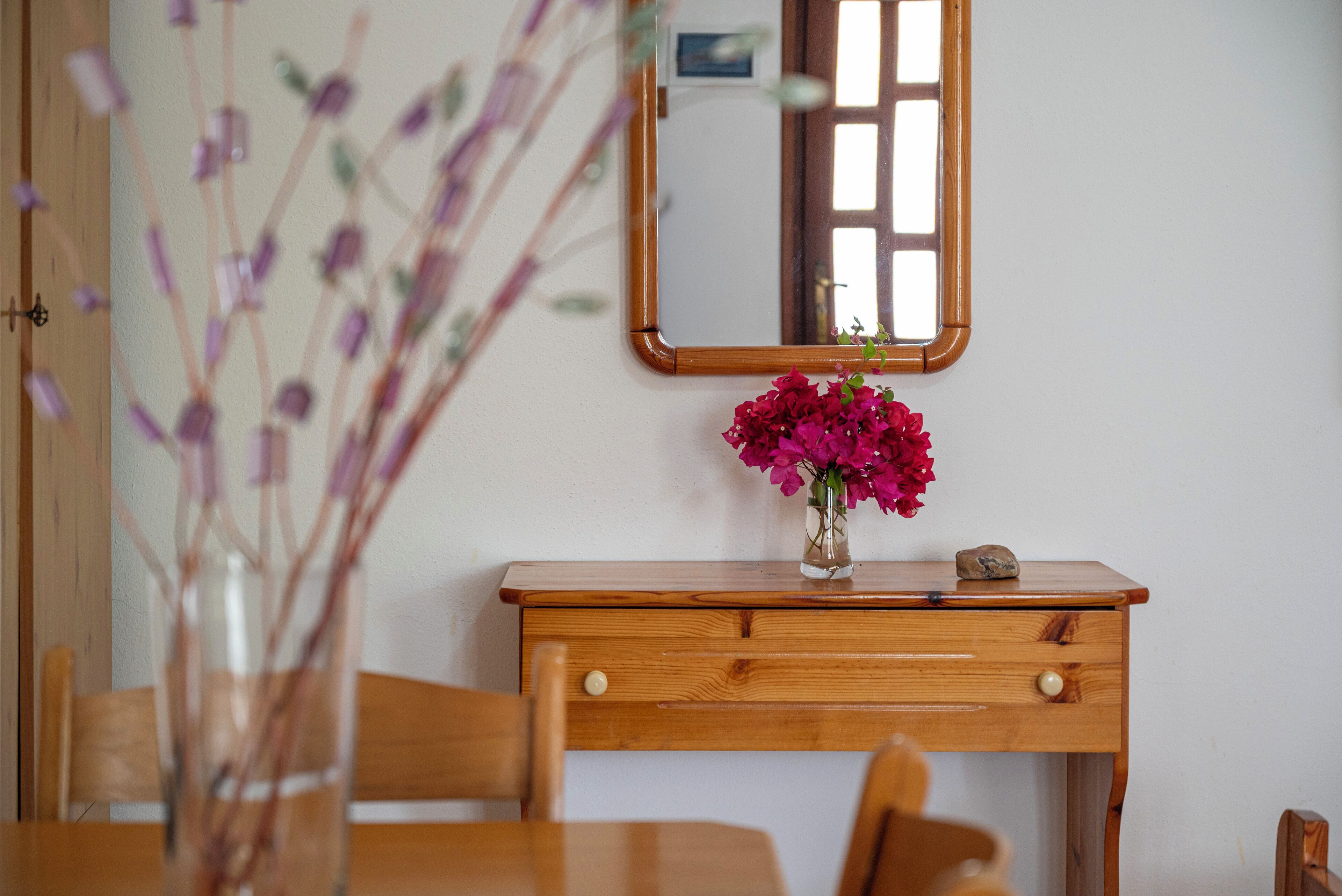 A wooden dresser with a mirror above it, a vase with flowers, and a wooden chair.